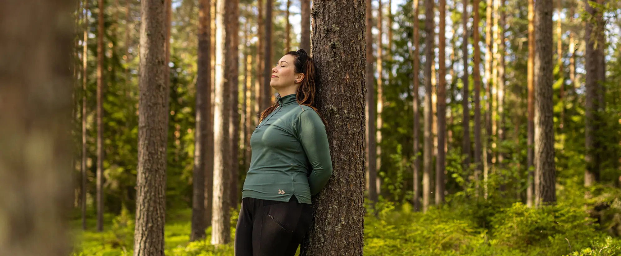 Metsänomistajanainen metsässä nojaamassa mäntyyn